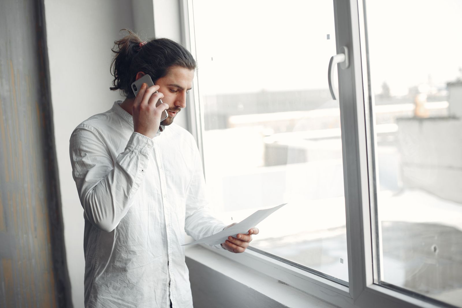 Man talking on a phone while reading a document by a window